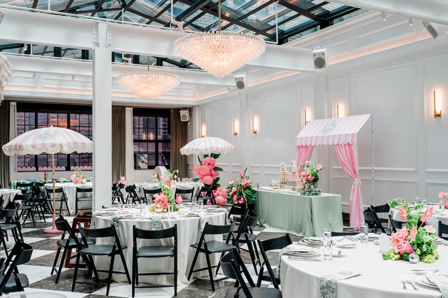 A room with skylights and a chandelier set for an event white round tables, pink flowers, and pink and white striped umbrellas.