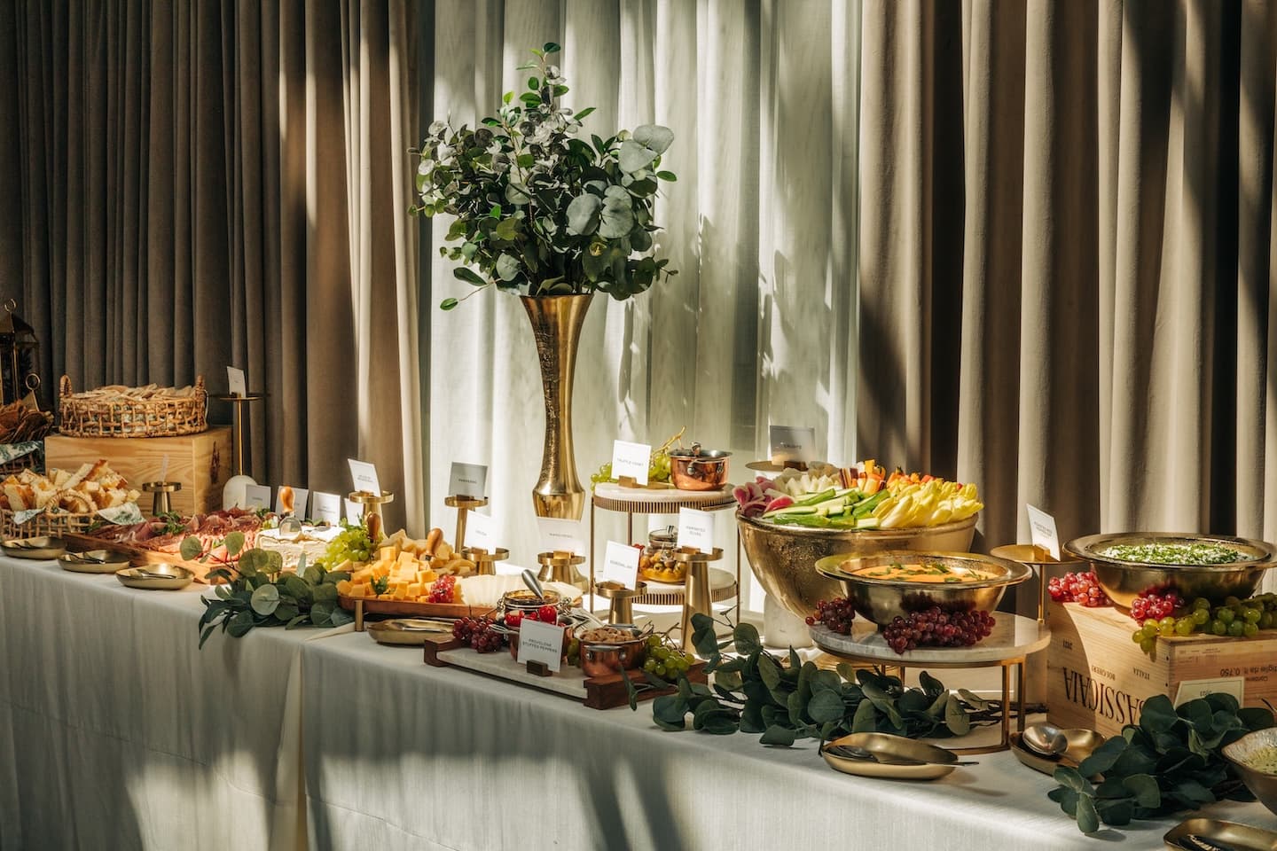 A long white table with charcuterie adorned with eucalyptus leaves. a brown and white curtain hangs behind it.
