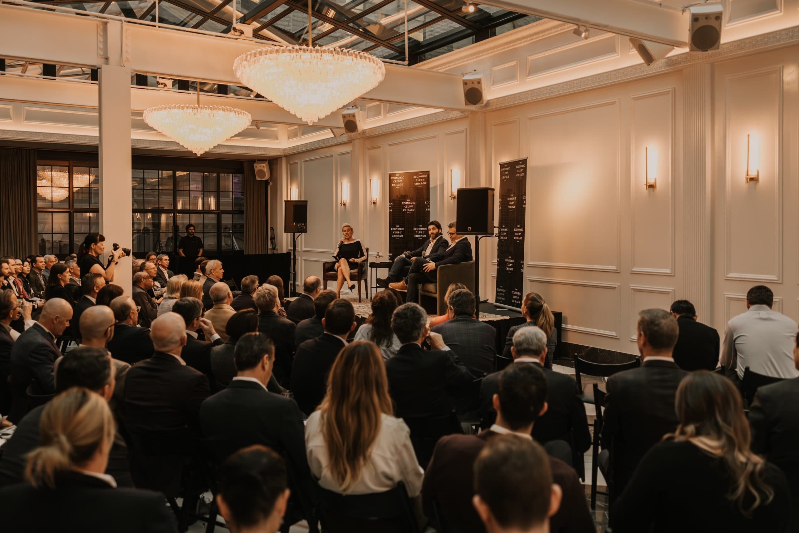 Meeting room filled with people in theater seating and seated lounge on stage for speakers.