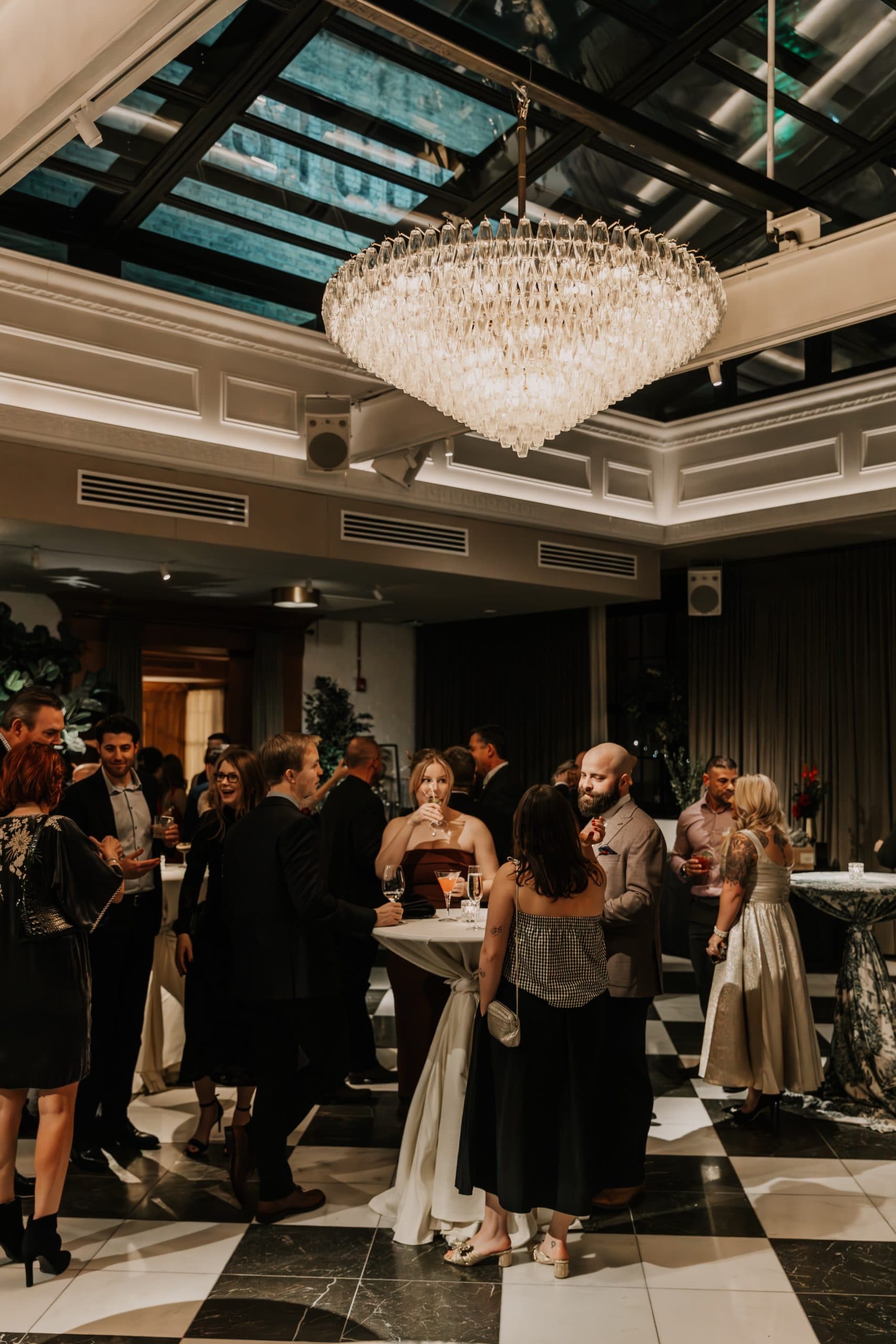 Room with black and white checkered floors, a chandelier hanging, and a skylight with people mingling.